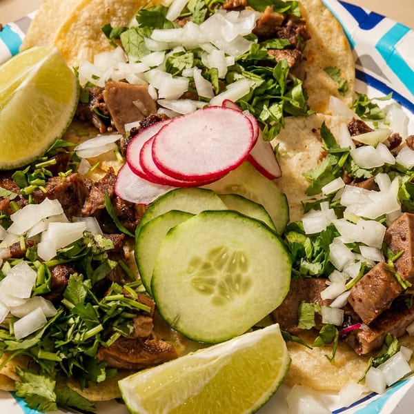 Three street tacos with meat, cilantro, onions, radishes, cucumbers, and lime wedges on a paper plate.