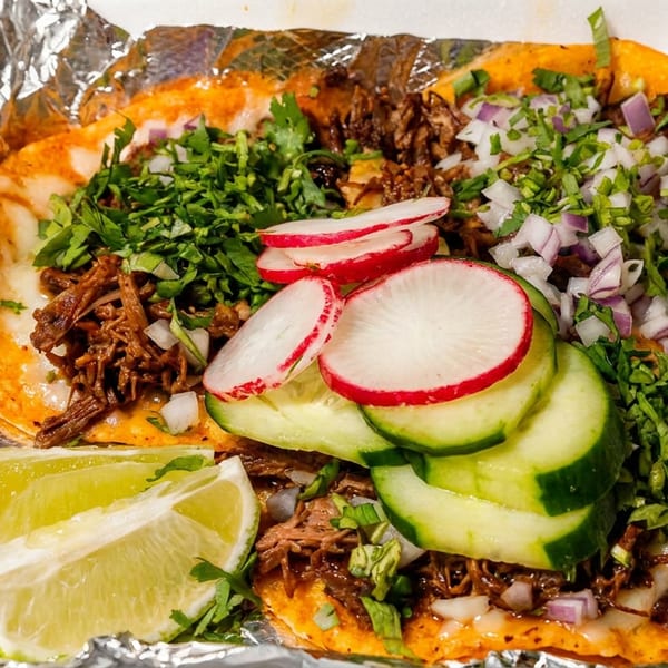 Three birria tacos with cilantro, onions, radishes, cucumbers, and lime wedges in foil.