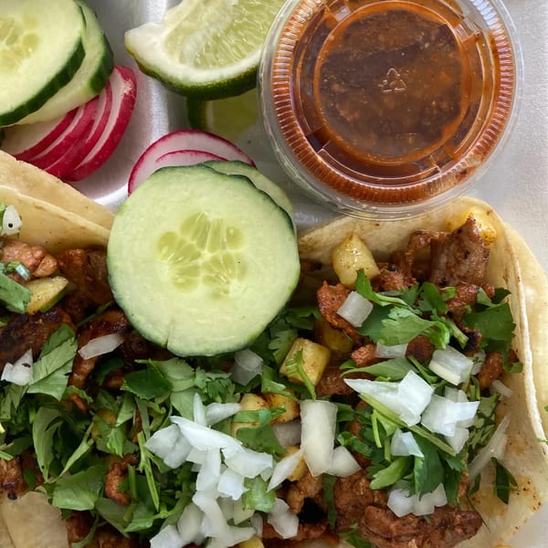 Two tacos with meat, cilantro, onions, and pineapple, served with lime, cucumber, radish, and salsa.