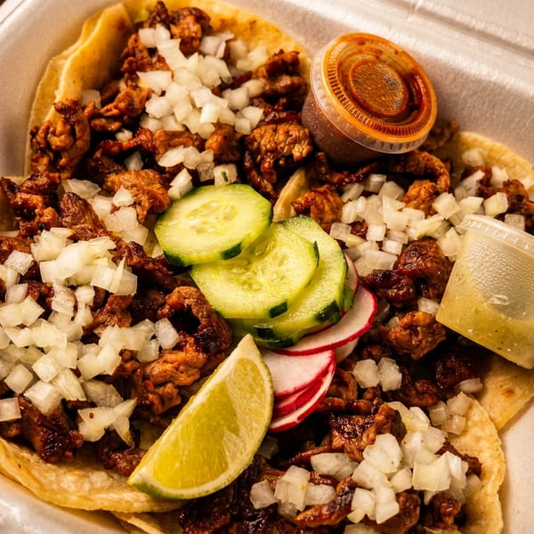 Three street tacos with meat, diced onions, cucumber, radish, lime, and two sauces in a to-go container.