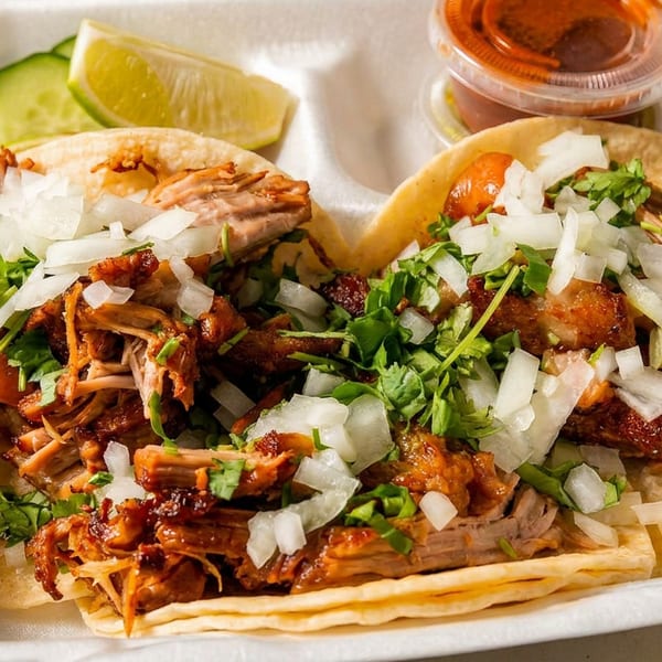 Two carnitas tacos topped with cilantro and diced onions, served with lime, cucumber, and salsa.